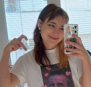A mirror selfie of me smiling, doing a heart symbol with my hand. My hair is split-dyed (brown and blonde). I'm wearing a t-shirt with a picture of Izzy from the tv show 'Our Flag Means Death'. A white wireless earbud is in my ear.