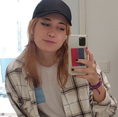 A mirror selfie of me with a black cap on. My phone case has the bisexual flag in it. I'm wearing a white flannel over a white t-shirt. My nails are painted black.