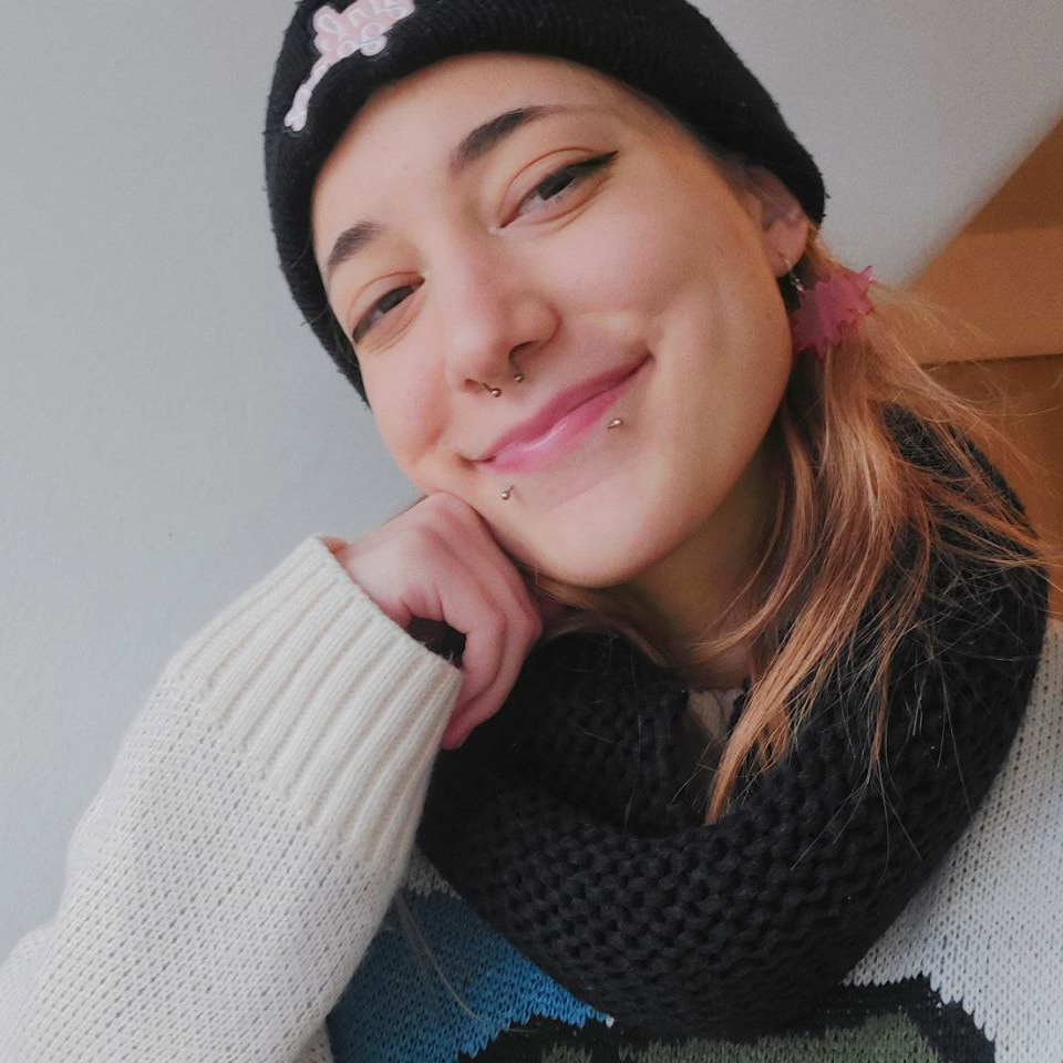 Me smiling into the camera, wearing a white sweater and a black beanie and scarf, and pink ear rings.